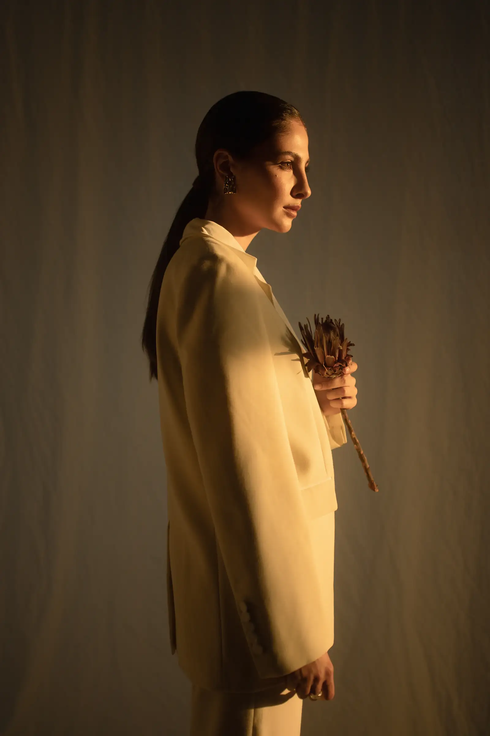 Woman in a camel blazer holding a dried flower, in profile against a warm dark wall