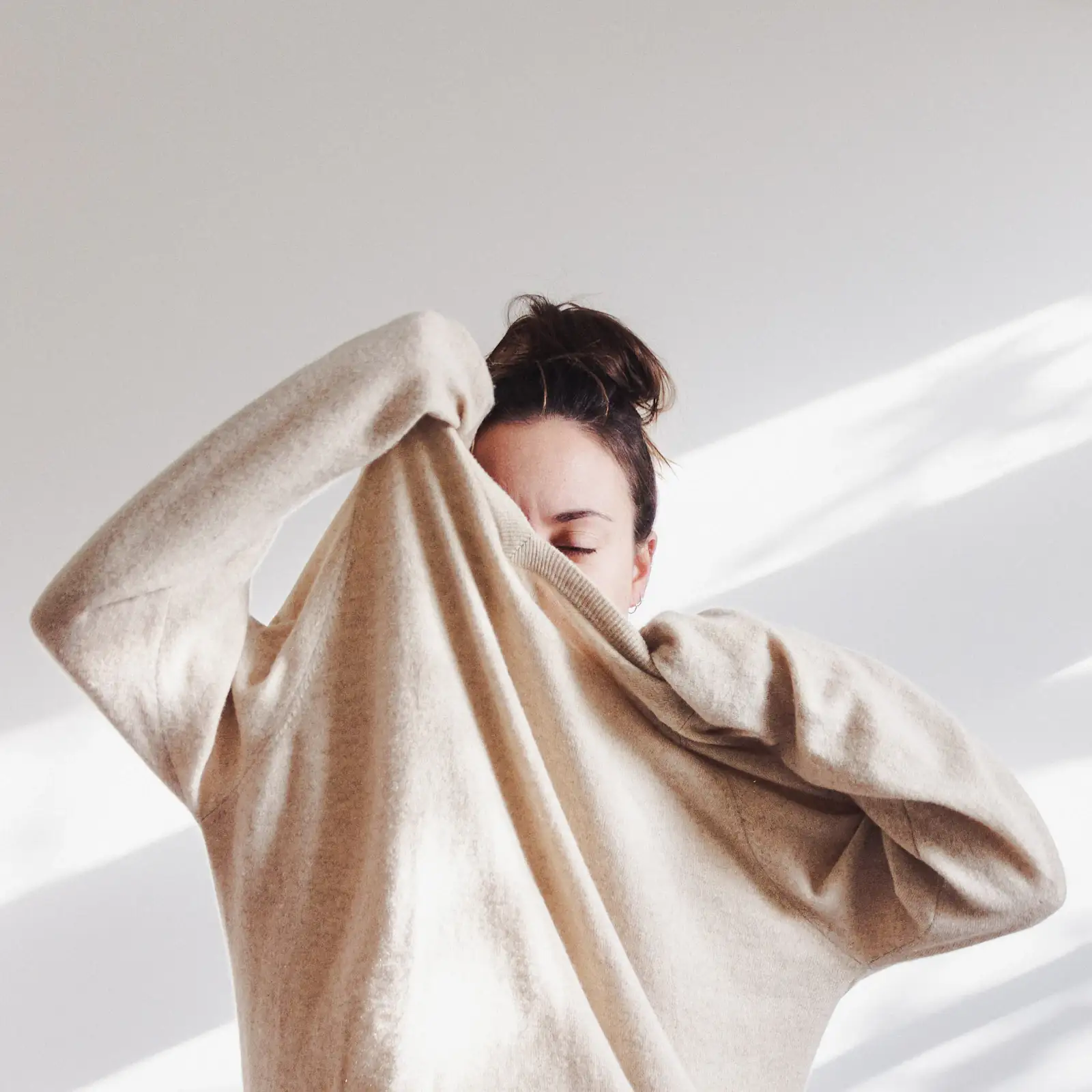 Woman in a cream knit pulling the sleeve over her face in soft natural light